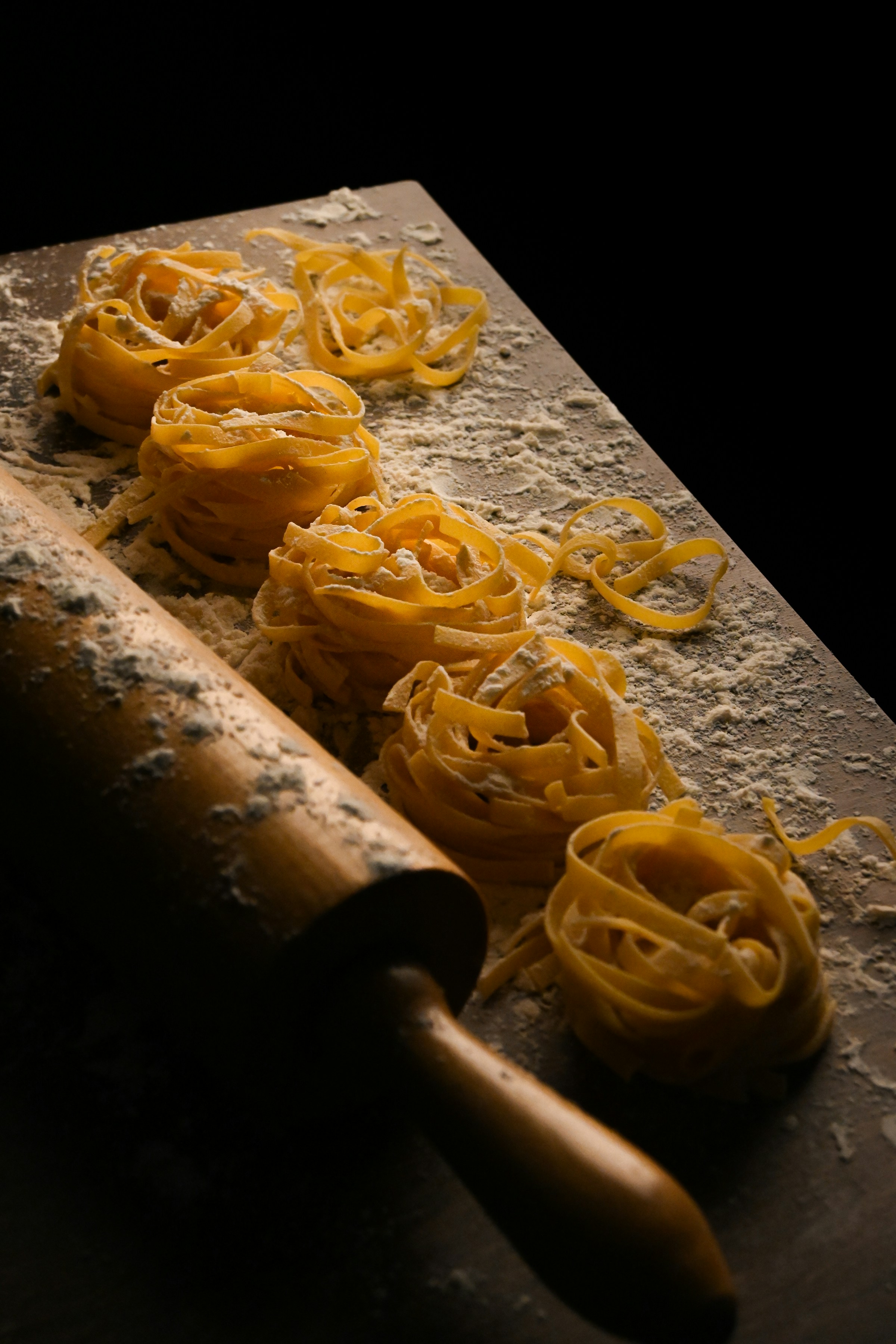 Fresh tagliatelle nests and a rolling pin on a flour-dusted work surface